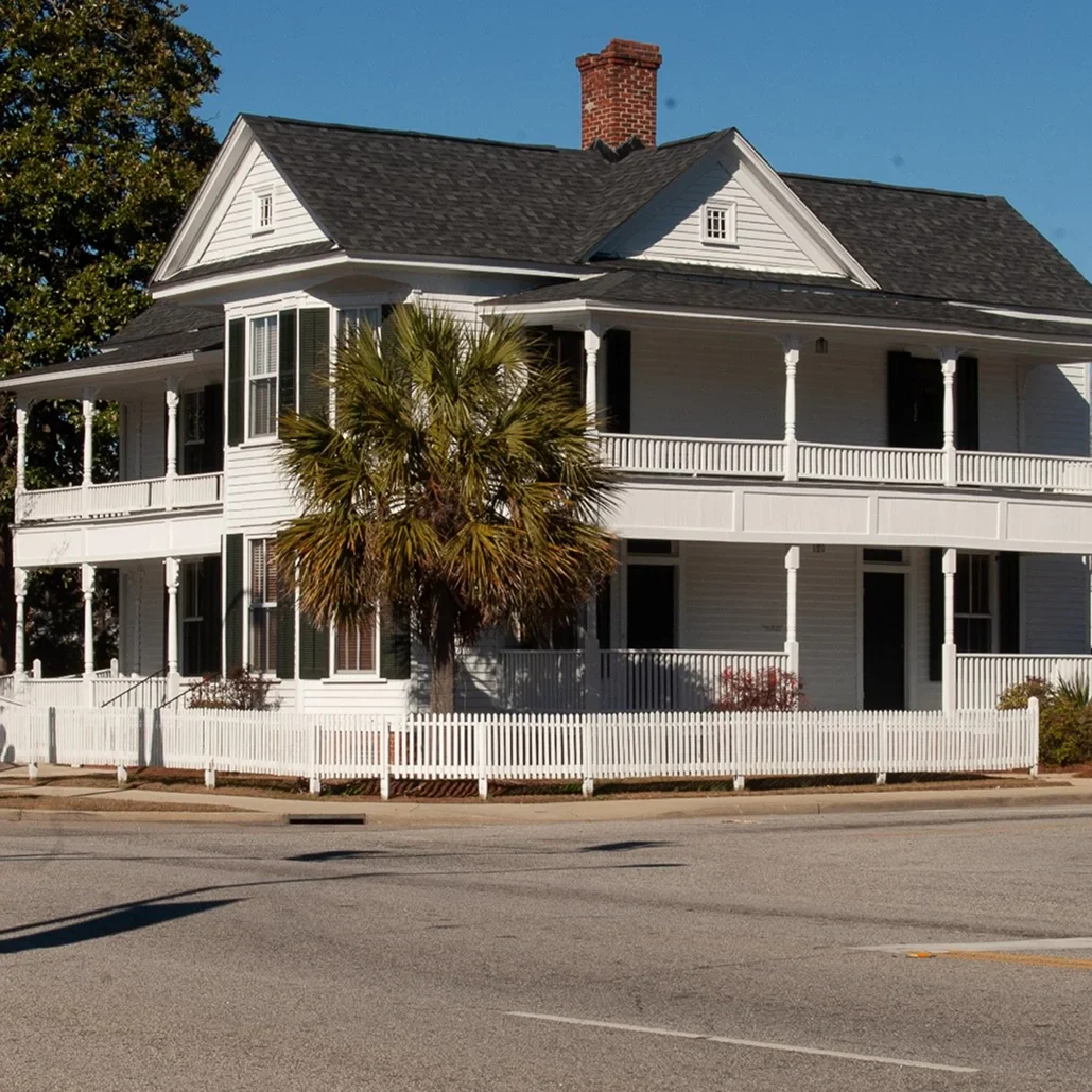 A charming white historic house with a gabled roof, palm tree, and picket fence, located at a street corner under a clear blue sky.