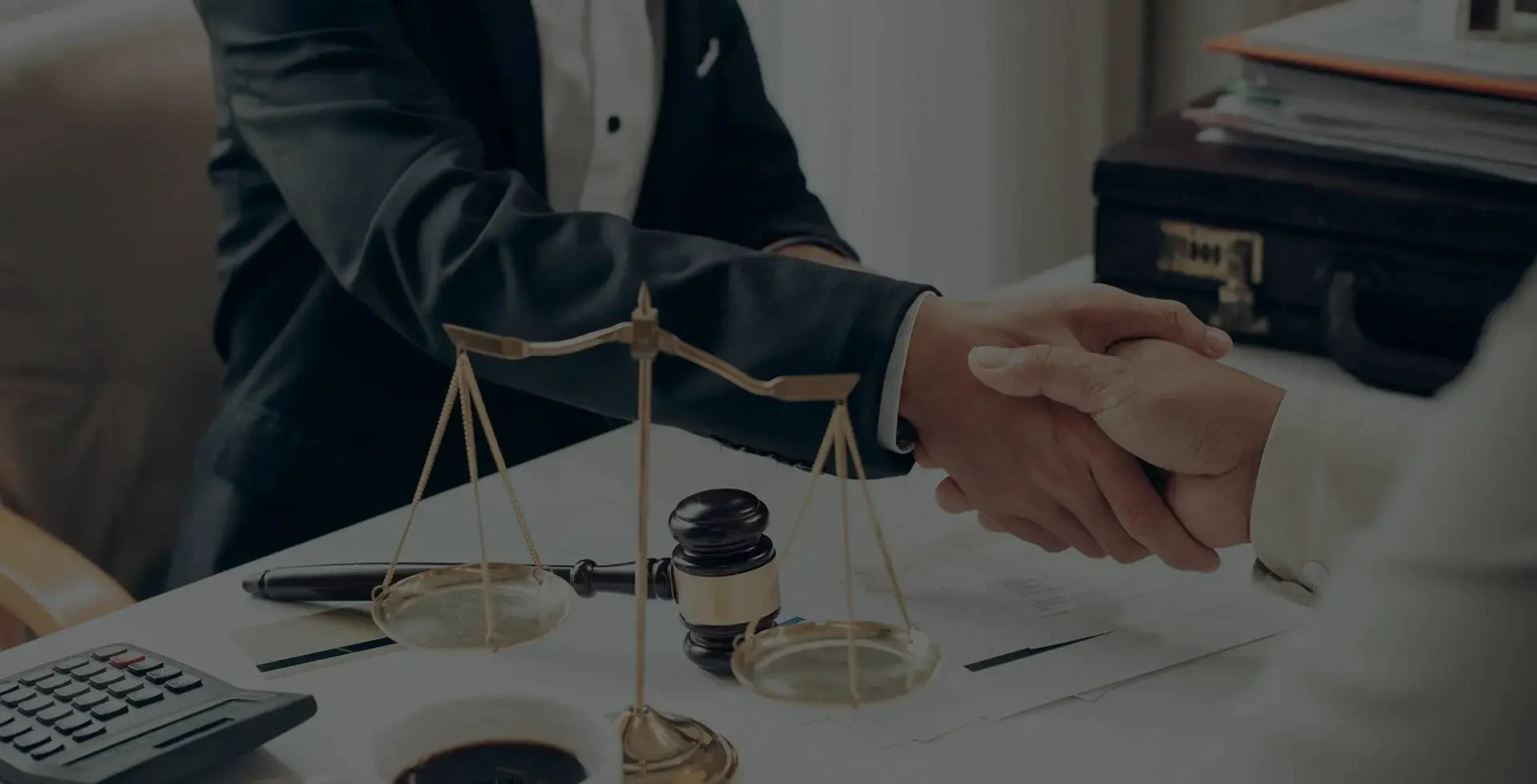 Two people shake hands over a desk with a gavel and scales of justice, symbolizing a legal agreement or partnership.