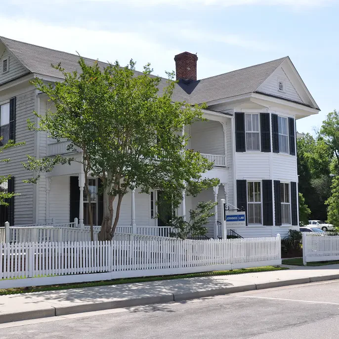 A charming two-story white house with black shutters, surrounded by a white picket fence and greenery, houses Jennings Law office.